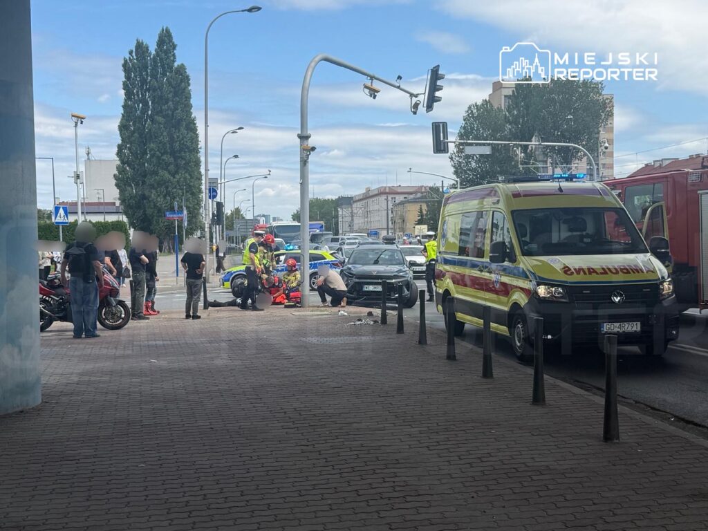 Grupa ludzi obserwuje interwencję ratunkową, podczas gdy strażacy udzielają pomocy poszkodowanemu na chodniku.