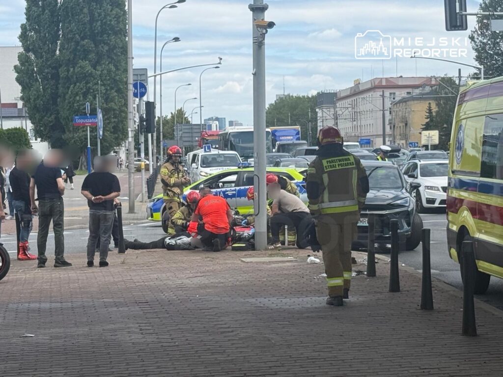 Grupa ratowników udziela pomocy poszkodowanemu na chodniku obok ruchliwej ulicy, z policyjnym radiowozem w tle.
