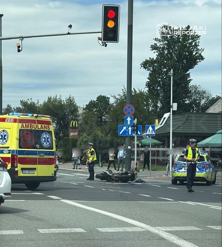 Ambulans oraz pojazdy policyjne na miejscu wypadku, obok leżącego motocykla na ruchliwej ulicy.