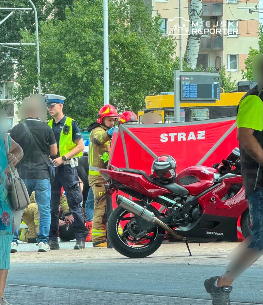 Grupa strażaków i policjantów udziela pomocy poszkodowanemu w wypadku motocyklowym na ruchliwej ulicy. W tle widoczny czerwony motocykl marki Honda.
