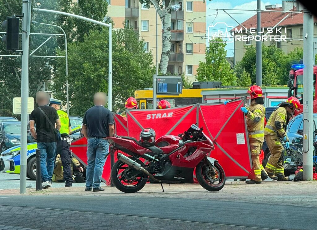 Grupa strażaków ustawia czerwoną osłonę wokół miejsca zdarzenia, podczas gdy policjant rozmawia z mężczyzną obok motocykla.