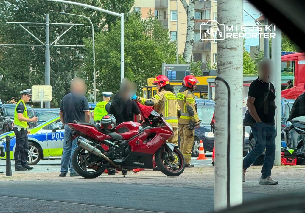 Grupa strażaków w hełmach rozmawia z poszkodowanym mężczyzną obok czerwonego motocykla, w tle policyjny radiowóz.