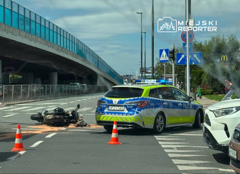 Motocykl leży na drodze, obok stoi radiowóz policyjny z włączonymi sygnałami świetlnymi.