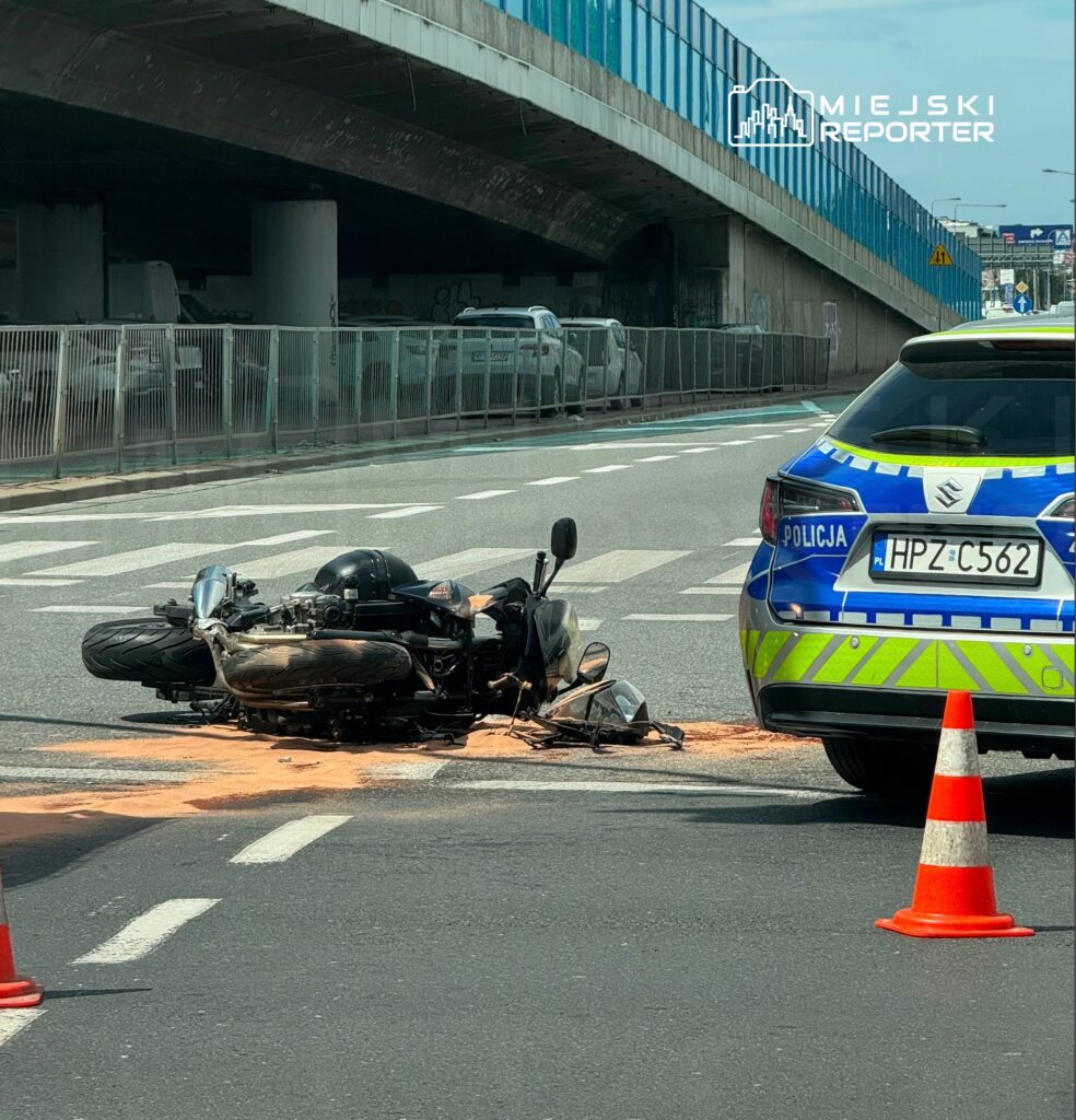 Motocykl leży na jezdni, obok stojący radiowóz policyjny oraz oznakowane pachołki. Na drodze widoczna plama cieczy.