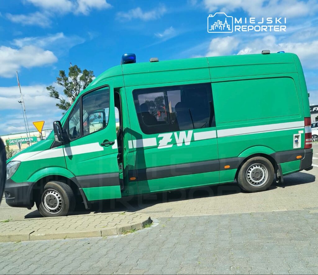 Zielony pojazd służbowy z napisem "ZW" zaparkowany na placu, w tle błękitne niebo z białymi chmurami.