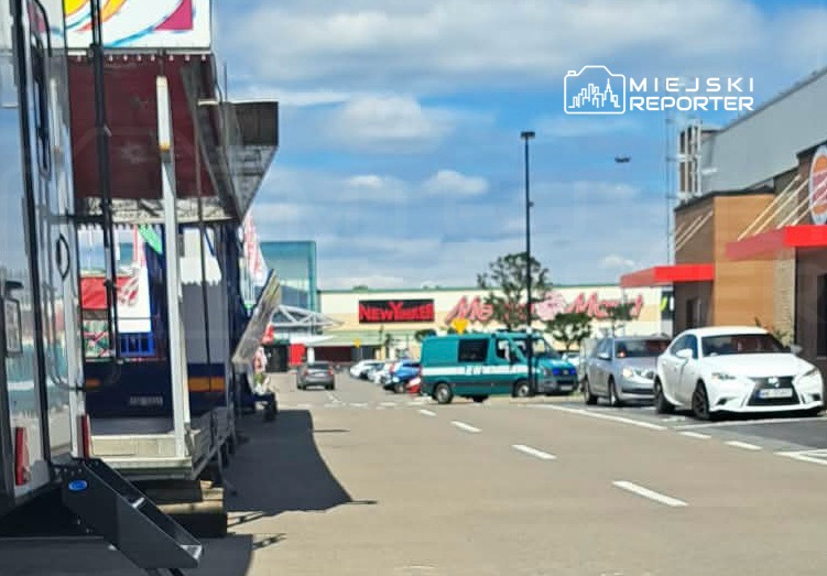 Ruchliwy parking przed centrum handlowym, z ustawionymi przy nim przyczepami oraz zaparkowanymi samochodami.