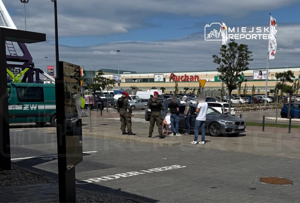 Grupa mężczyzn rozmawia na parkingu przed hipermarketem, obok stoi radiowóz i pojazd służbowy.