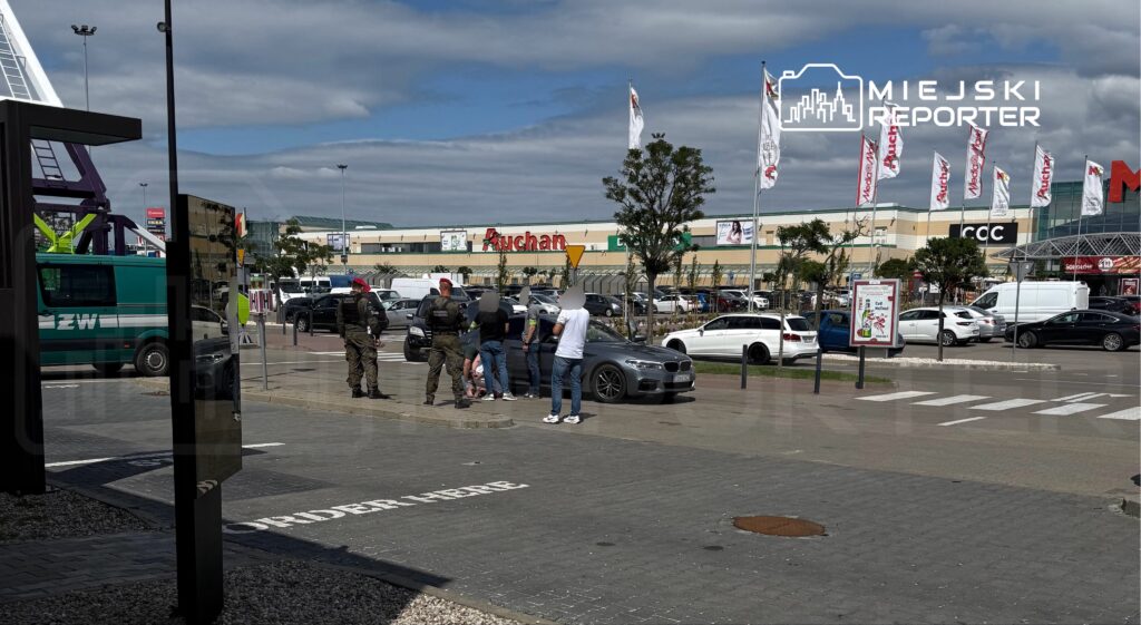 Grupa osób, w tym dwóch mężczyzn w mundurach, rozmawia na parkingu przed centrum handlowym.