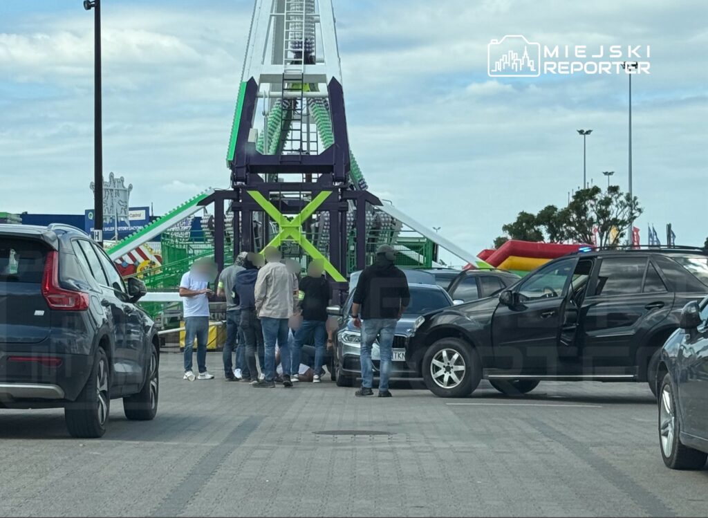 Grupa osób stoi wokół samochodu na parkingu, w tle widać atrakcje wesołego miasteczka.