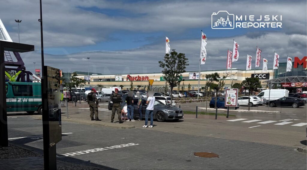 Grupa osób stoi obok samochodu w parkingu przed dużym centrum handlowym, w tle widoczne flagi i budynki.