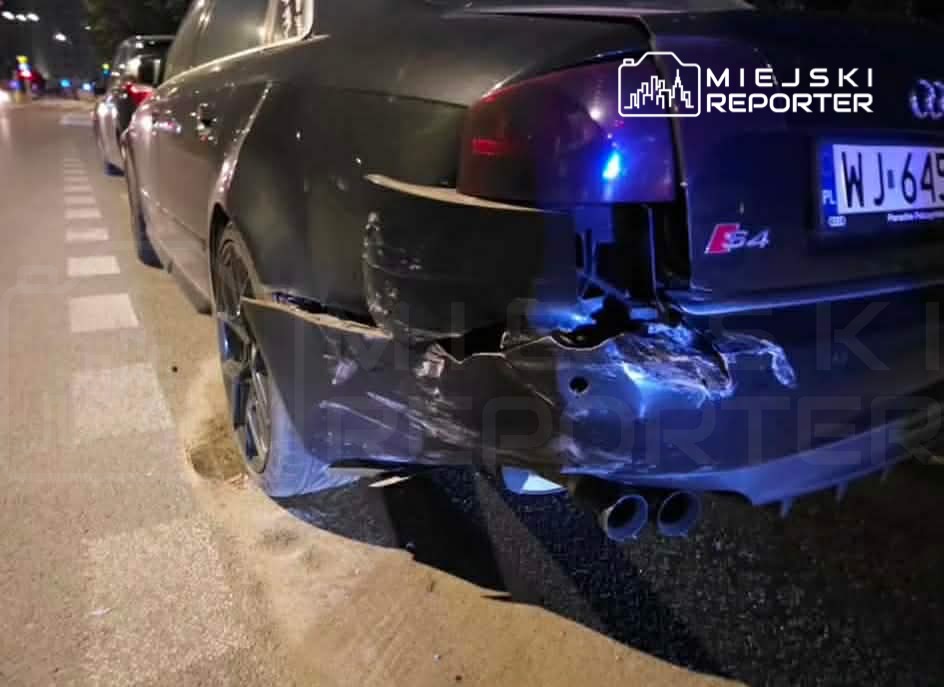Uszkodzony czarny samochód marki Audi z wgniecioną tylną częścią stoi na ulicy nocą.