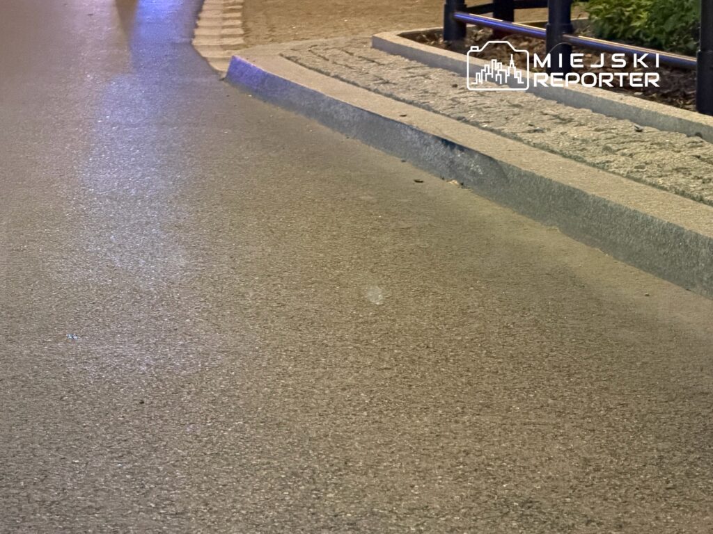 Nocna ulica z mokrym asfaltem odbijającym światła, z krawężnikiem i elementami małej architektury.