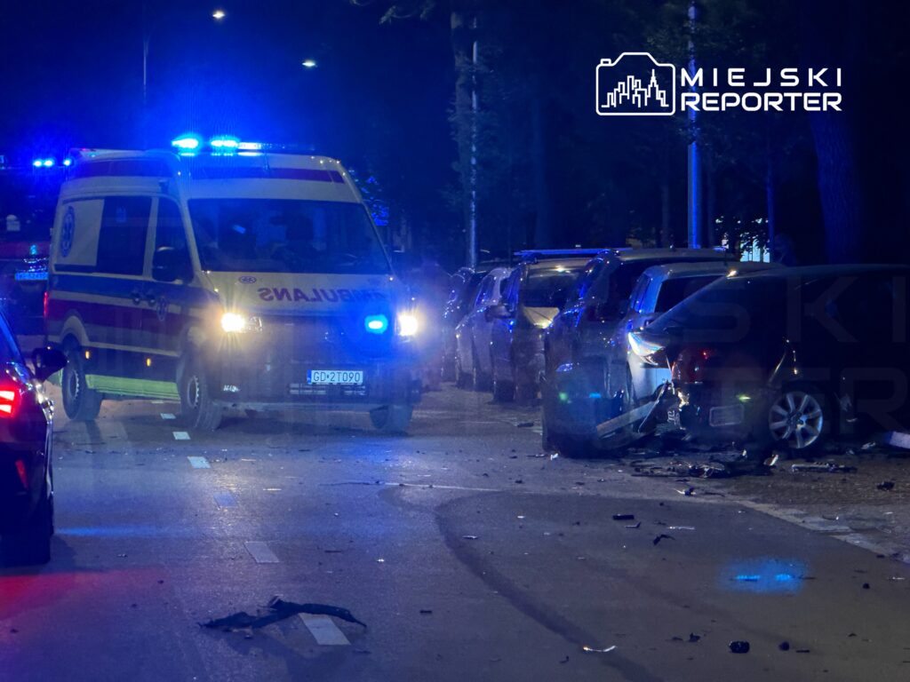 Ambulans z włączonymi sygnałami świetlnymi stoi na ulicy obok zderzonych samochodów, wokół leżą fragmenty karoserii.