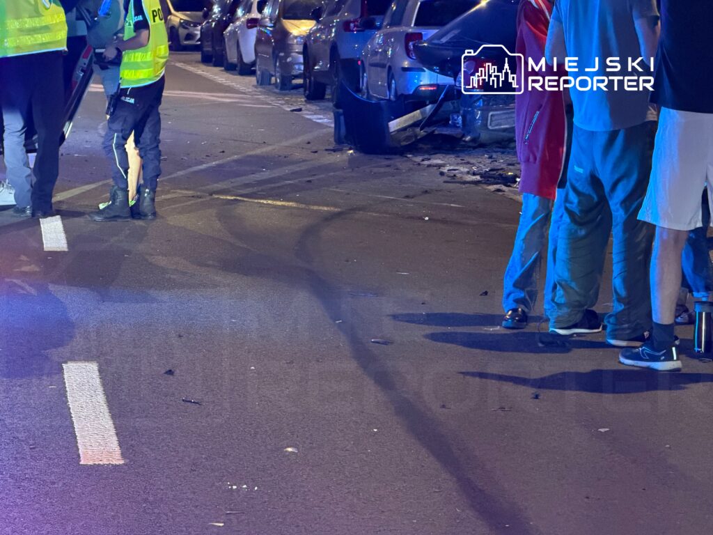 Grupa osób w odblaskowych kamizelkach rozmawia z policjantami na oświetlonej ulicy, obok uszkodzonych samochodów.