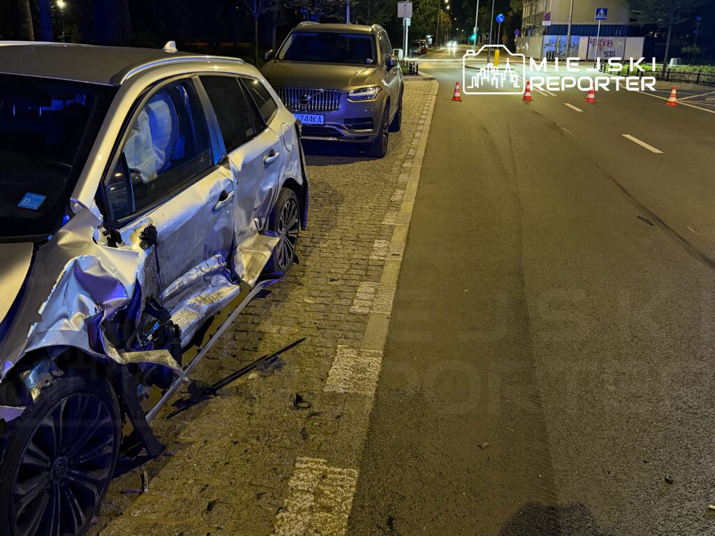 Uszkodzony samochód osobowy z wgniecioną boczną stroną zaparkowany na ulicy, obok stoją inne pojazdy.