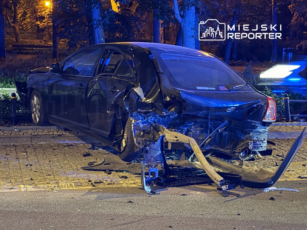 Czarny samochód osobowy z uszkodzeniami tyłu stoi na chodniku w oświetlonym parku, obok leżą fragmenty zderzaka.