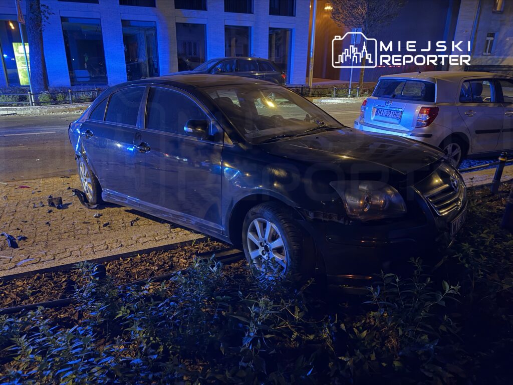Czarny samochód osobowy zaparkowany na chodniku, obok widać zniszczoną nawierzchnię i nieoznakowane pojazdy w tle.