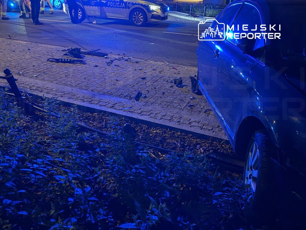 Na jezdni leżą fragmenty szkła, a w tle policjanci prowadzą czynności na miejscu zdarzenia.
