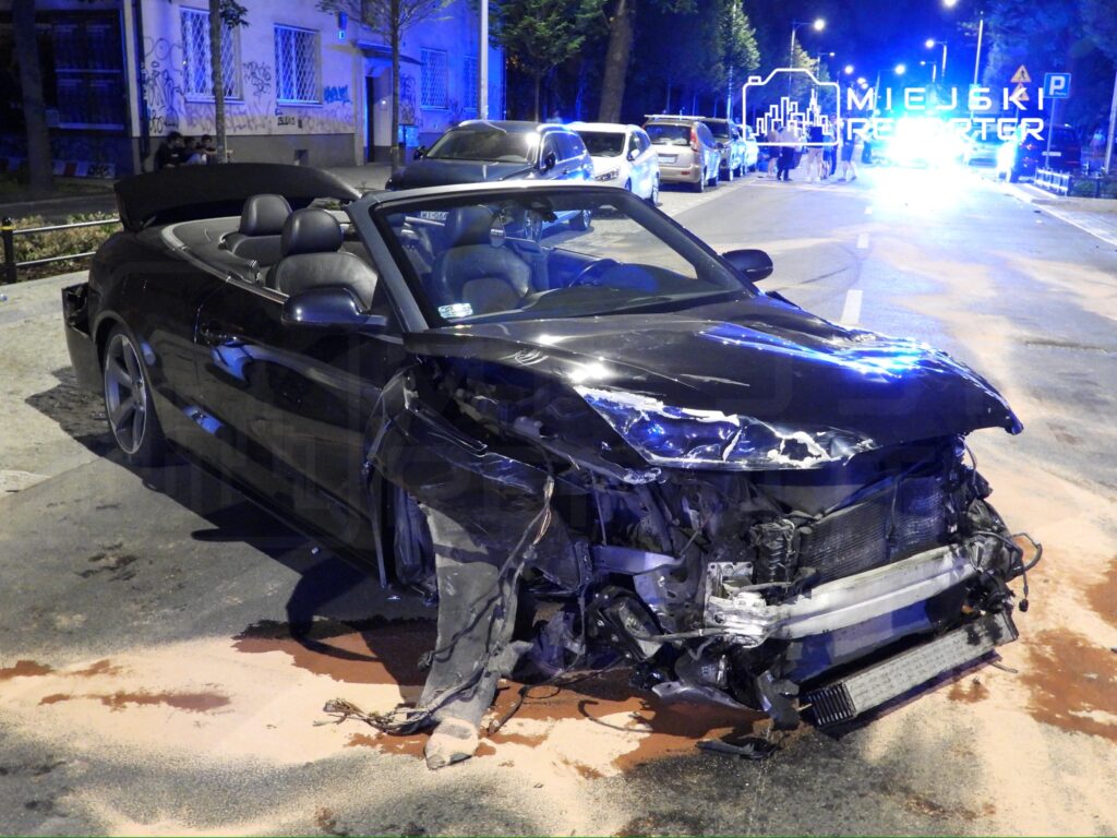 Uszkodzony czarny kabriolet stoi na ulicy, obok widać policyjne światła i grupę ludzi obserwujących zdarzenie.