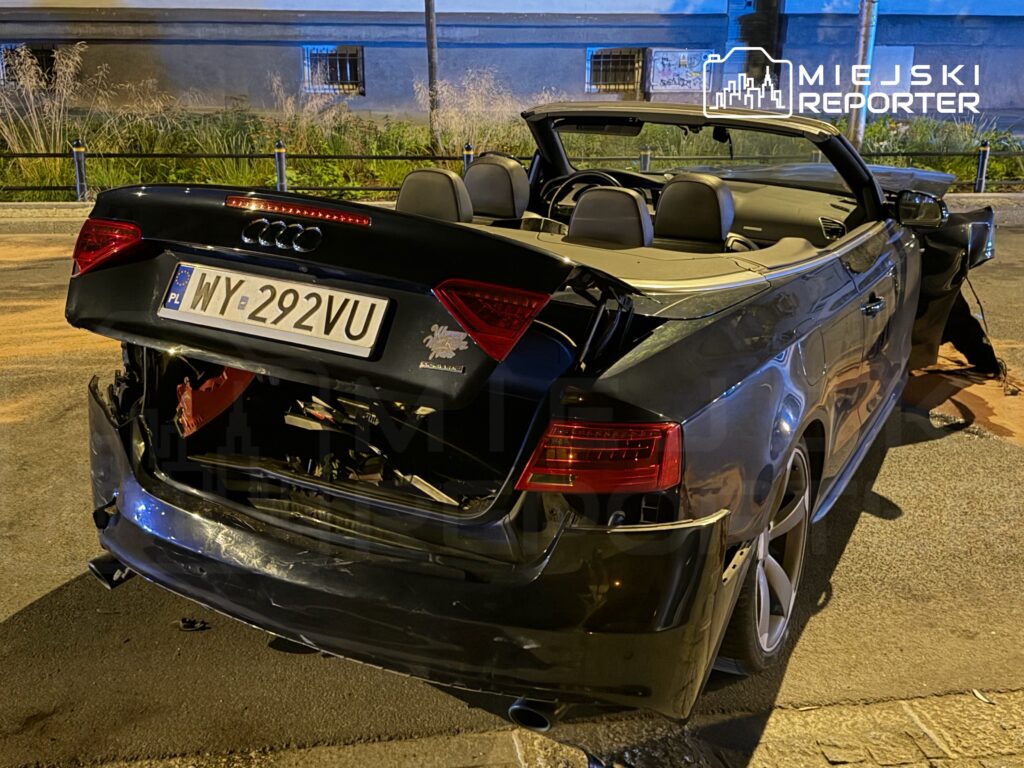 Uszkodzony czarny kabriolet Audi stoi na ulicy, z widocznym uszkodzeniem tyłu i otwartym dachem.