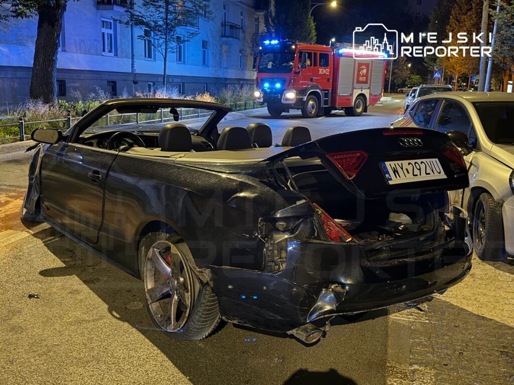 Uszkodzony kabriolet zaparkowany na ulicy, z widoczną otwartą klapą bagażnika, obok przejeżdżający wóz strażacki.
