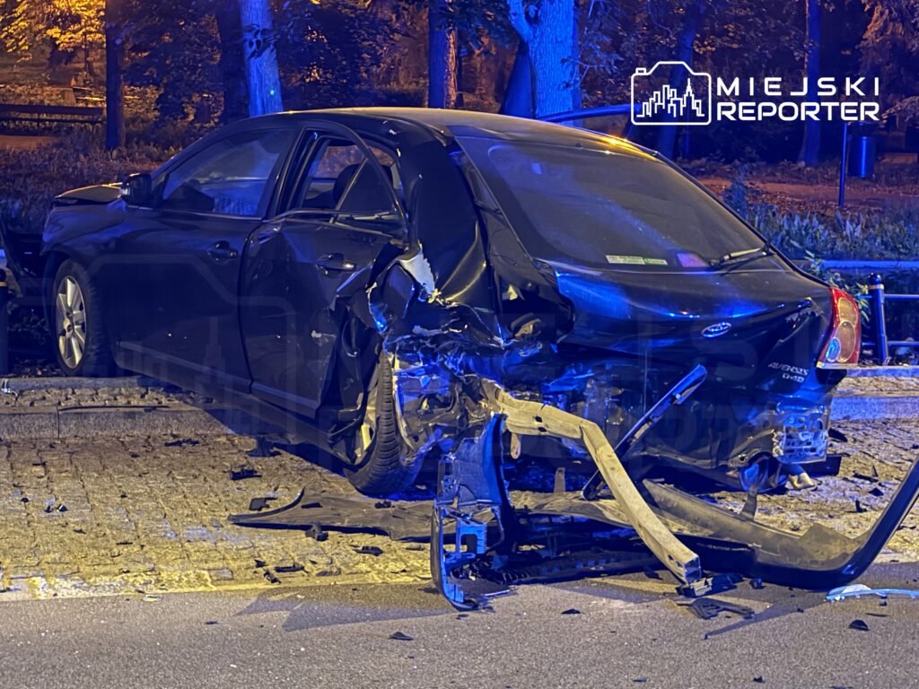 Zniszczony czarny samochód osobowy leży na poboczu, z jednym kołem w powietrzu, otoczony odłamkami.