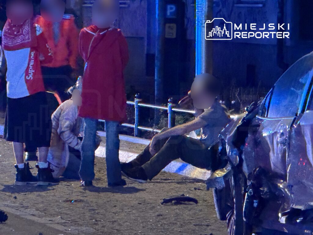 Grupa osób w casualowych ubraniach zbiera się wokół poszkodowanego mężczyzny siedzącego na ulicy obok zniszczonego samochodu.