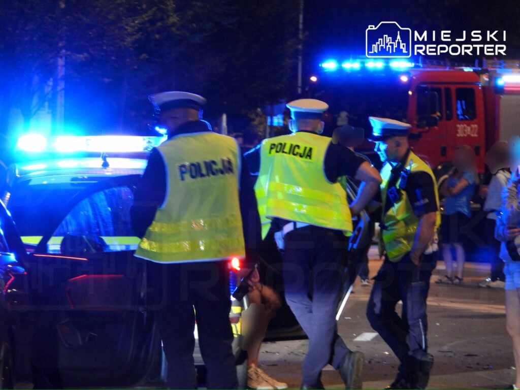 Grupa policjantów w odblaskowych kamizelkach prowadzi działania przy radiowozie z włączonymi sygnałami świetlnymi.