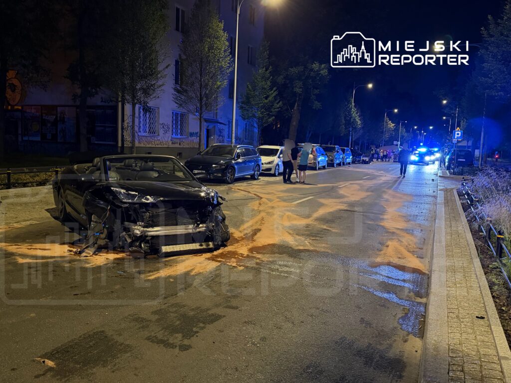 Uszkodzony czarny kabriolet stoi na ulicy, obok grupy ludzi i jadących samochodów, w nocy przy oświetleniu ulicznym.