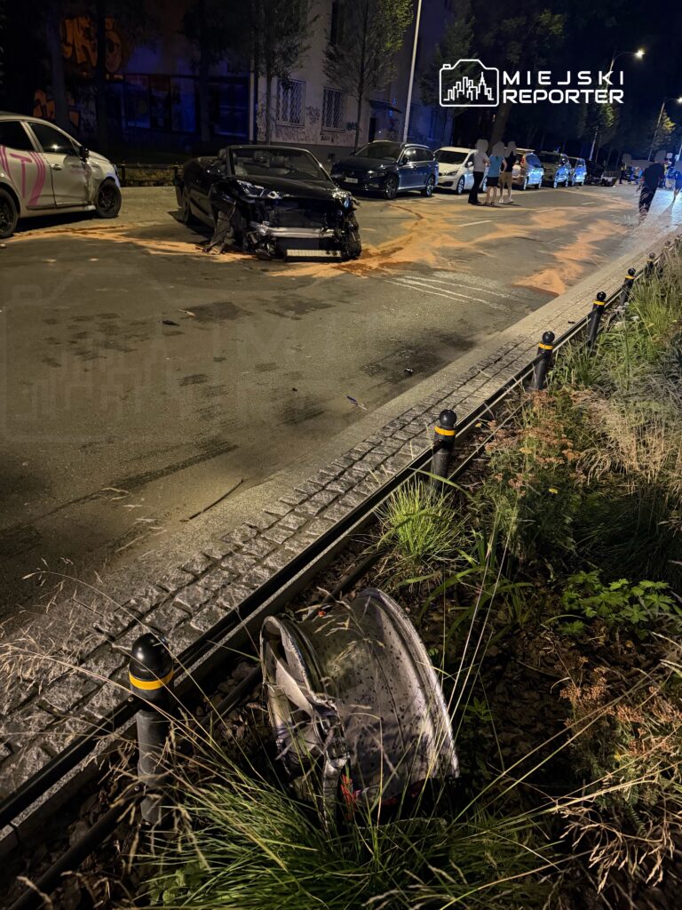 Uszkodzony czarny samochód sportowy stoi na poboczu ulicy, obok leży zdemolowane koło, a w tle widać inne pojazdy.