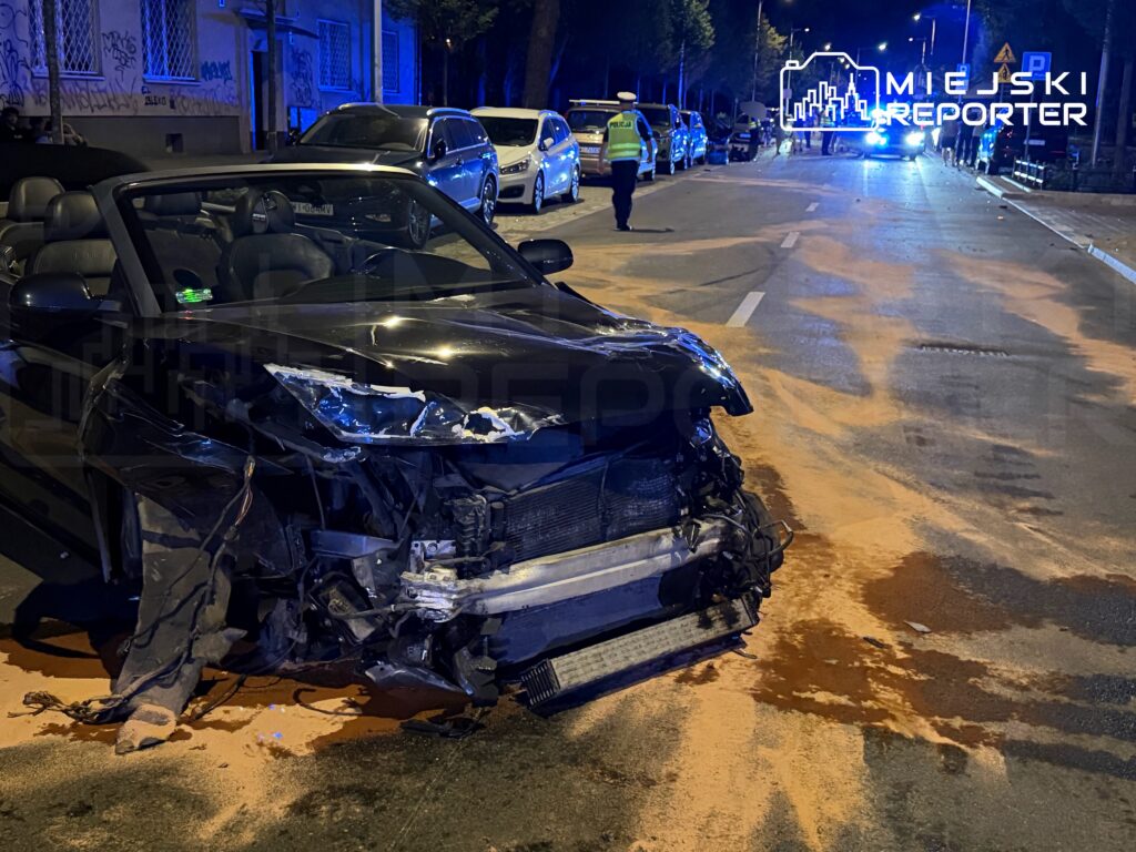 Czarny kabriolet z uszkodzoną przednią częścią stoi na ulicy, obok widać policjanta oraz rozlany piasek.