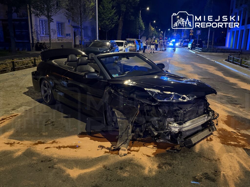 Czarny kabriolet z uszkodzoną przednią częścią stoi na drodze, wokół widać policyjne światła i grupę ludzi.