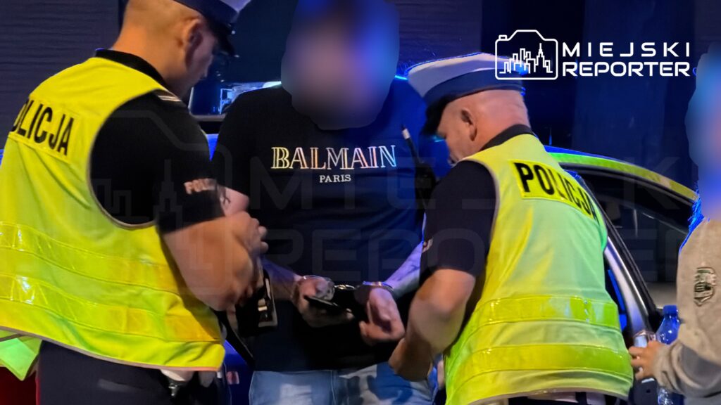 Mężczyzna w czarnej koszulce z napisem "BALMAIN" przekazuje dokumenty dwóm policjantom w kamizelkach odblaskowych.