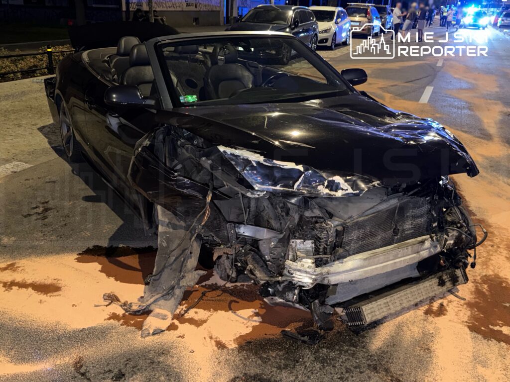 Czarny kabriolet z uszkodzoną przednią częścią stoi na drodze, wokół widać pojazdy policyjne i zbierających się gapiów.