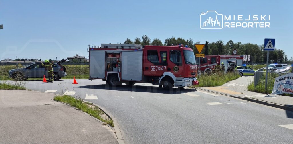 Strażak kieruje ruchem na skrzyżowaniu, obok zaparkowany wóz strażacki i radiowóz policyjny. W tle widać budynki.