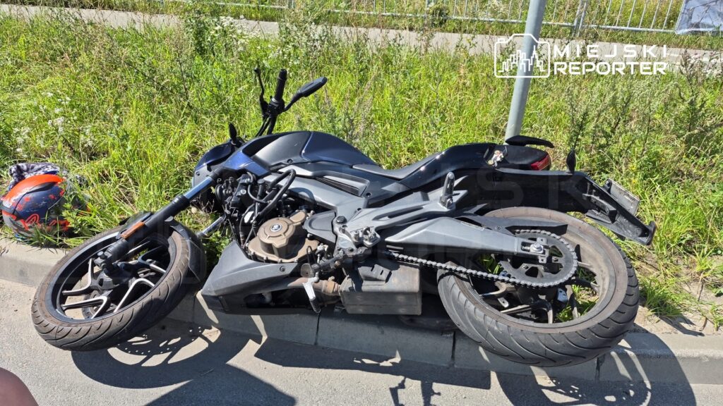 Czarny motocykl leży na boku obok drogi, a obok niego znajduje się kask. W tle widać trawę i ogrodzenie.
