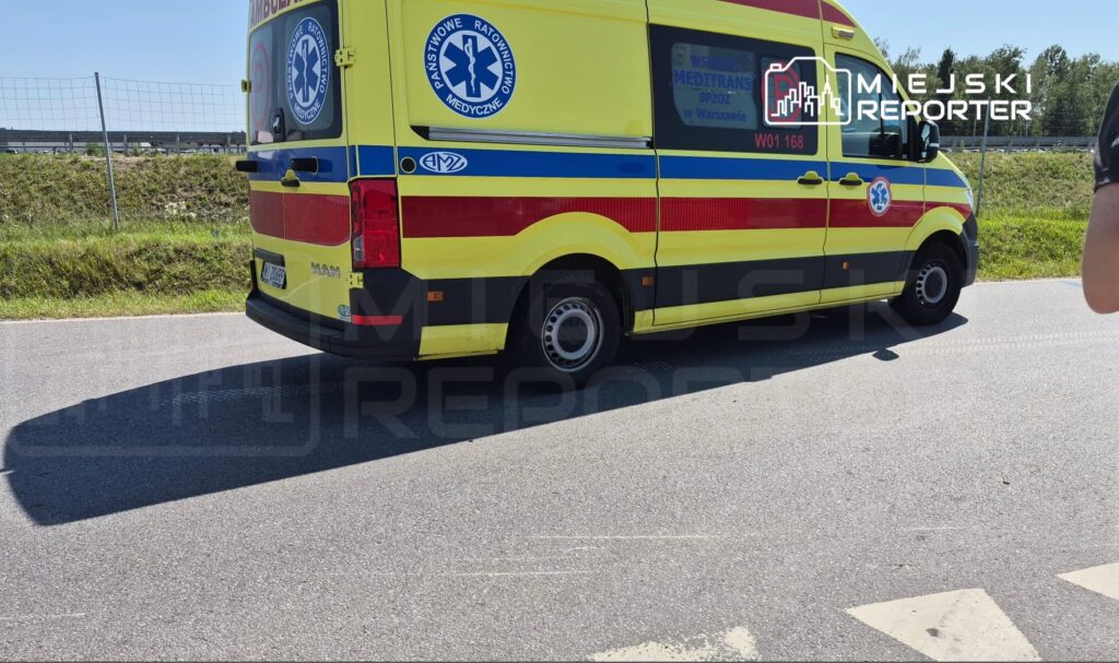 Karetka pogotowia w żółto-czerwonym kolorze stoi na poboczu drogi, obok zielonej trawy i ogrodzenia.
