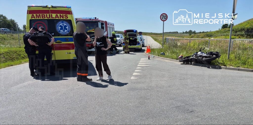 Na drodze stoi karetka, obok której dwaj policjanci rozmawiają z kobietą, a w tle widać przewrócony motocykl.