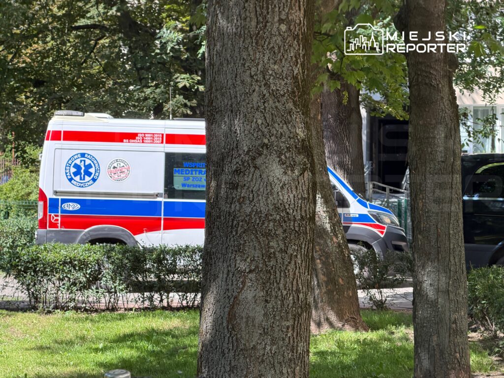 Karetka pogotowia z niebiesko-czerwonymi oznaczeniami stoi na ulicy w otoczeniu drzew i krzewów.