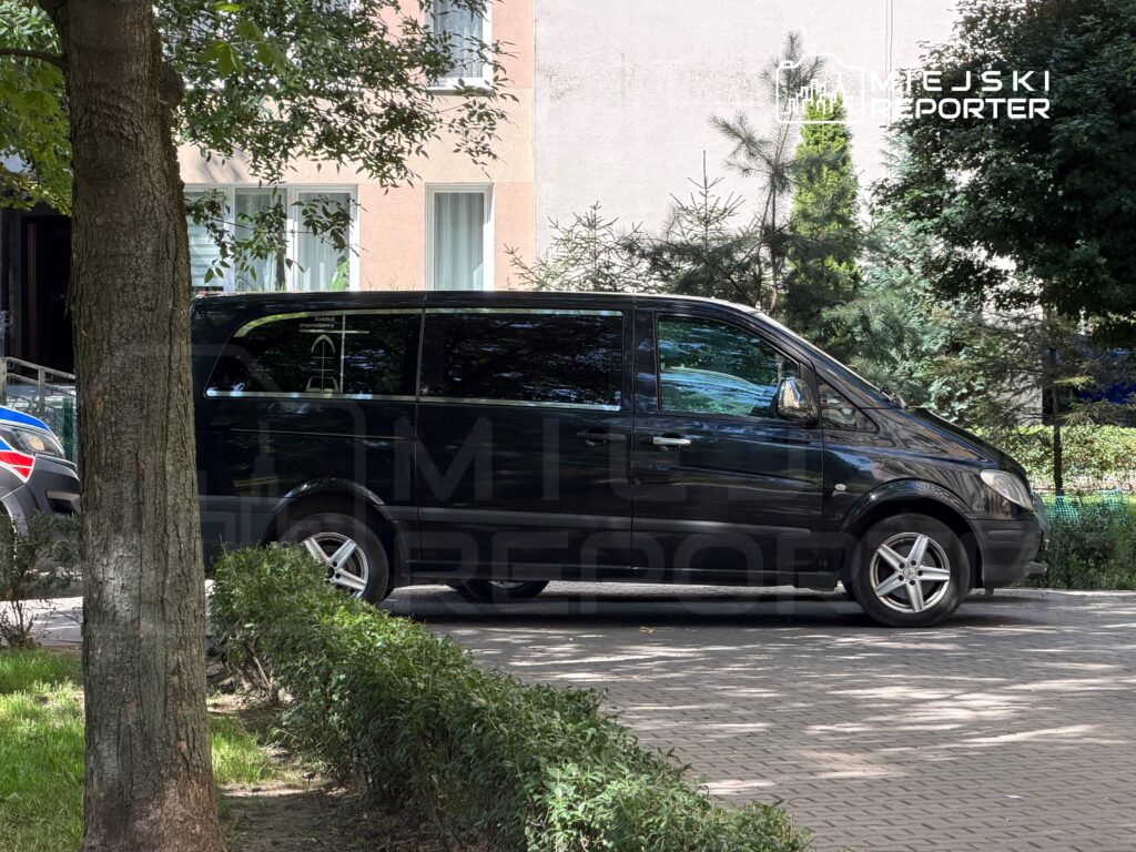 Czarny van zaparkowany na chodniku obok zielonych krzewów w miejskim otoczeniu, w tle widoczne budynki mieszkalne.