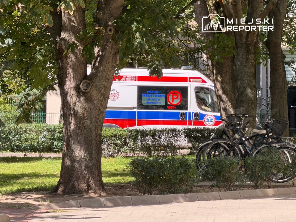 Karetka pogotowia parkowana obok drzew w miejskim parku, w tle rowery stojące na stojaku.