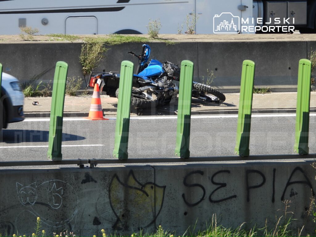 Motocykl leży na boku obok czerwonego pachołka na poboczu drogi, a w tle przejeżdża samochód.