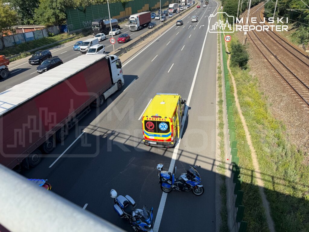 Karetka pogotowia przejeżdża obok stojących samochodów na ruchliwej drodze, obok torów kolejowych.