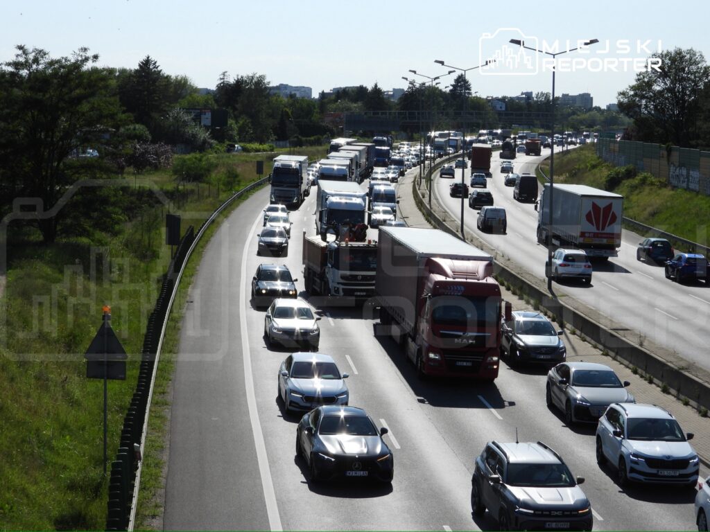 Ruch samochodowy na wielopasmowej drodze, z ciężarówkami i osobówkami w kierunku lewym, w otoczeniu zieleni.
