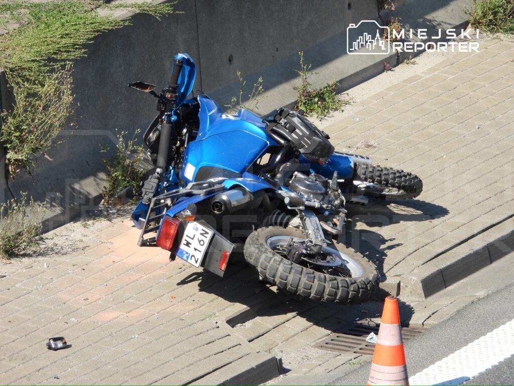 Niebieski motocykl leży na boku na chodniku obok betonowej ściany, zniszczony, obok pomarańczowy pachołek.