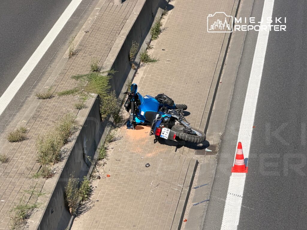 Motocykl w niebieskim kolorze leży na boku na poboczu drogi, obok znajduje się pomarańczowy trójkątny znak ostrzegawczy.