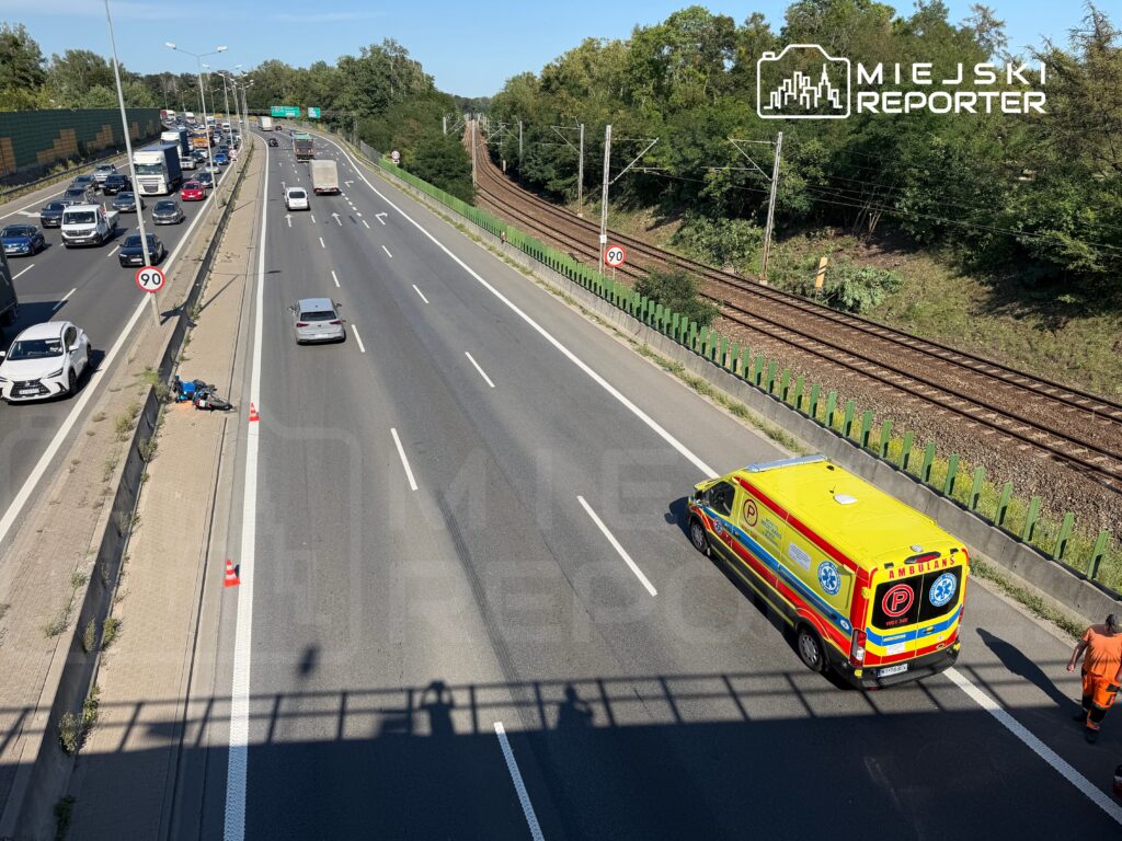 Na pasie drogi leżący rower oraz trójkąt ostrzegawczy, obok przejeżdża ambulans. W tle widoczny ruch samochodowy.