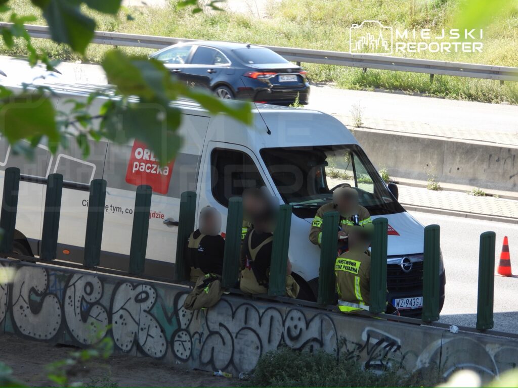Kierowca dostawczego samochodu rozmawia z ratownikami na poboczu drogi, obok graffiti i ustawionych pachołków.
