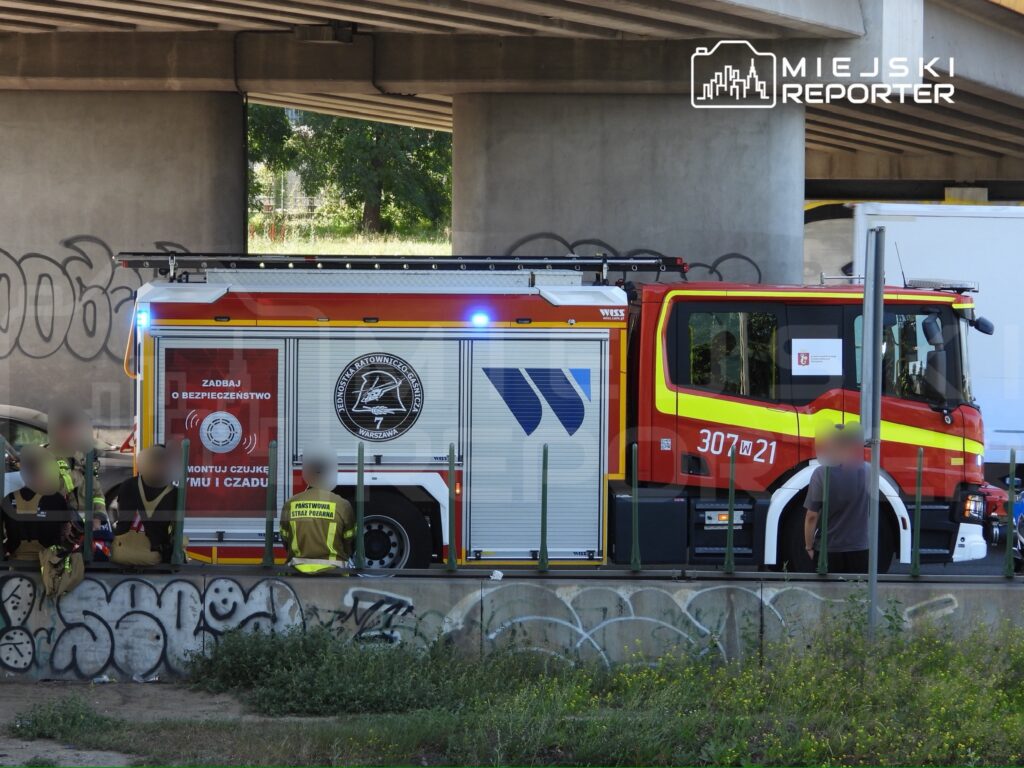 Wóz strażacki z oznaczeniem "307 W 21" zaparkowany pod mostem, z grupą strażaków w mundurach na tle graffiti.