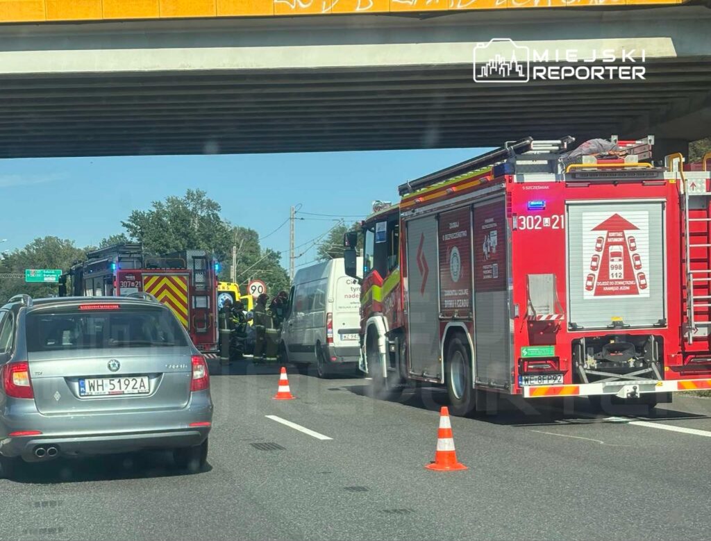 Na drodze stoi kilka wozów strażackich, a strażacy prowadzą działania przy białym vanie, otoczonym pachołkami.